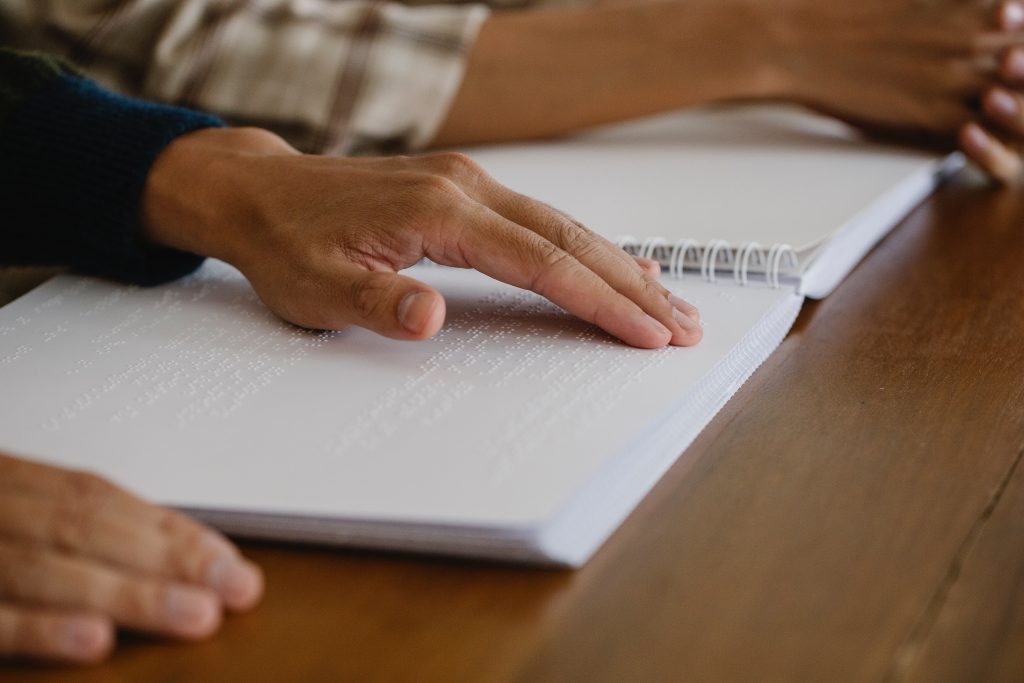 Hands reading a braille booklet. To their left, another pair of hands, as if the person to the reader’s left is there for assistance.